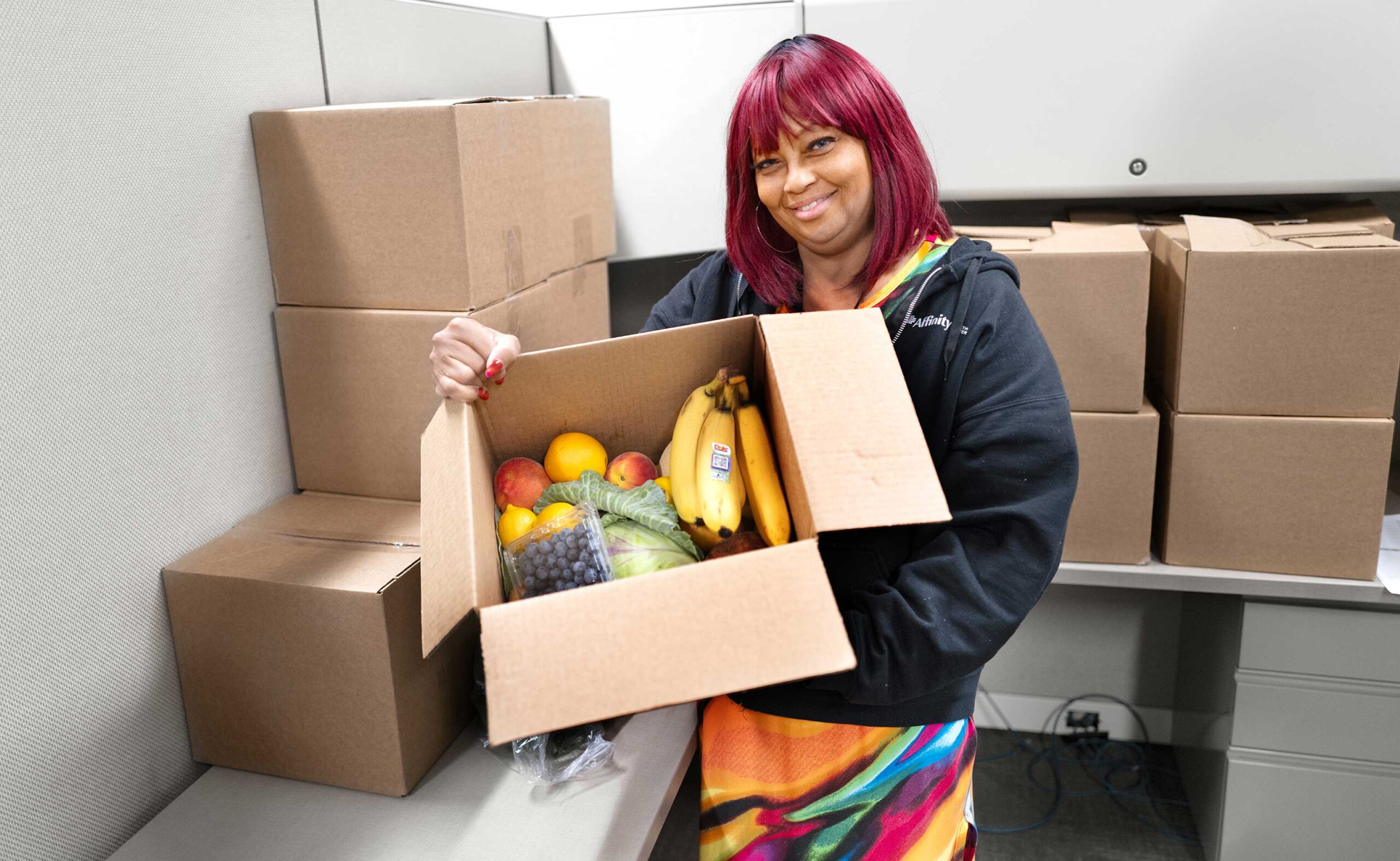 Community Health Worker holds a cardboard box filled with colorful fresh fruits and vegetables.
